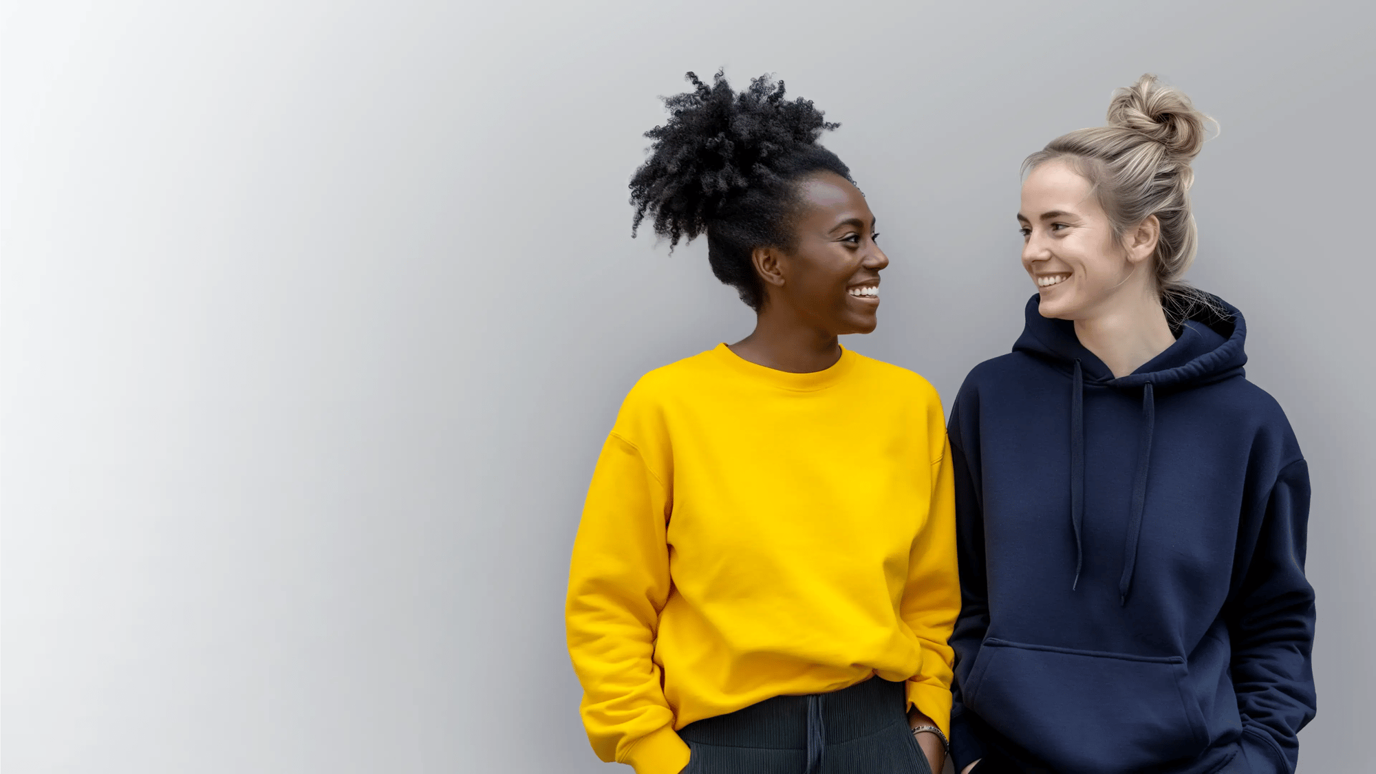 Two women in hoodies smiling at each other against a light background